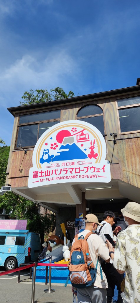 Entrance to the Mt. Fuji Panoramic Ropeway, featuring a colorful sign with a mountain graphic and visitors waiting in line.