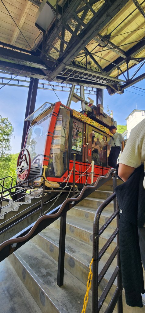 A red and black cable car station with a cable car waiting at the platform, surrounded by stairs and passengers.