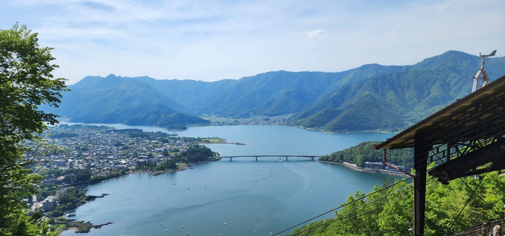 A panoramic view of a lake surrounded by mountains, with a town visible in the foreground and a bridge crossing the lake under a clear blue sky.