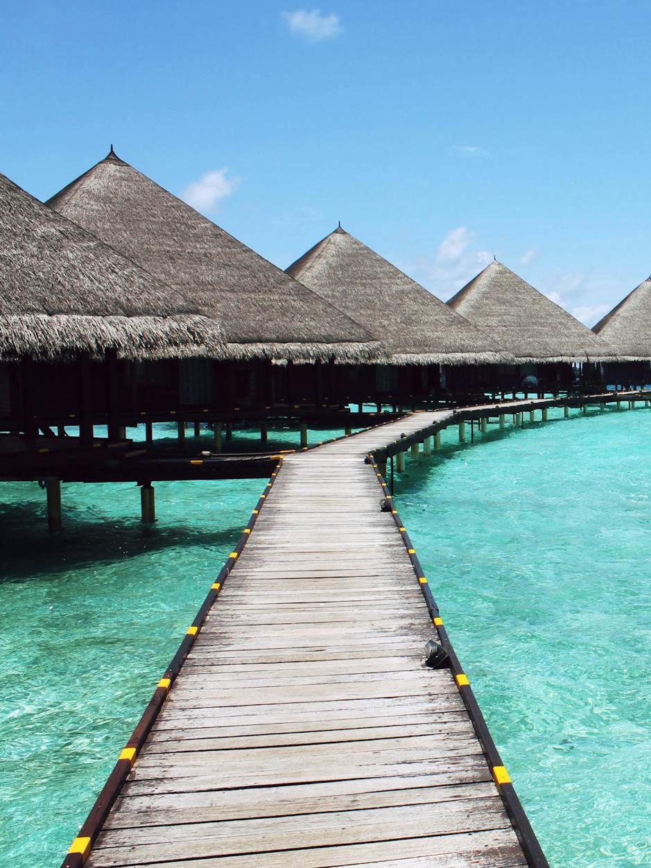 A wooden walkway leading to thatched-roof overwater bungalows surrounded by clear turquoise water under a blue sky.