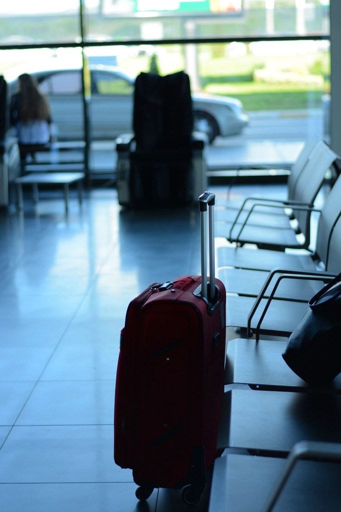 A red suitcase on wheels stands in an airport seating area, with blurred figures and a window in the background.
