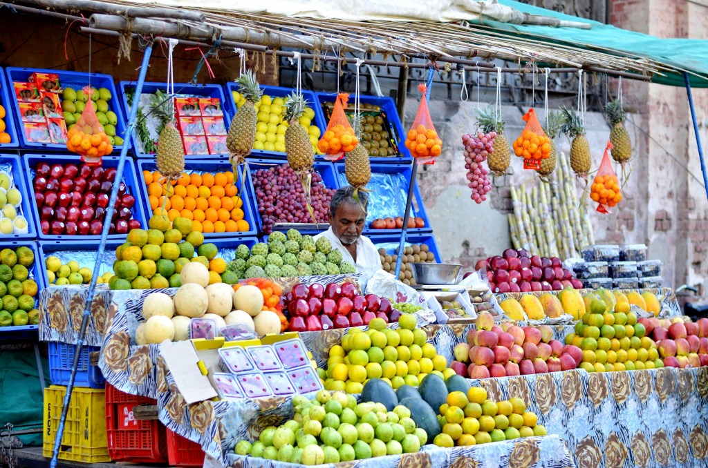 A fruit vendor at a market stand selling a variety of fresh fruits including pineapples, oranges, apples, melons, and more, with colorful displays and baskets.