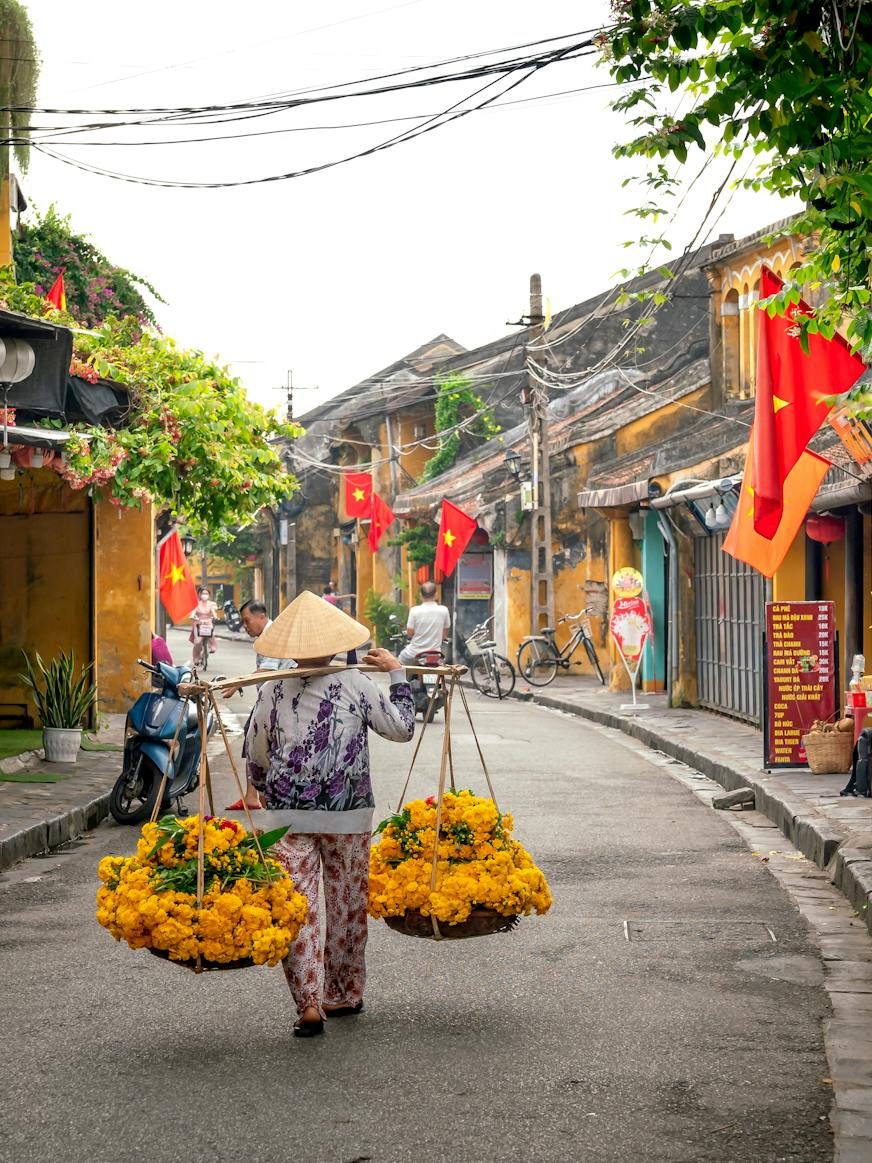 A street scene in Hoi An, Vietnam, featuring a woman in traditional attire carrying two baskets of yellow flowers. The background shows colorful buildings adorned with greenery and Vietnamese flags. Sidewalks are lined with potted plants and there are a few bicycles parked nearby.