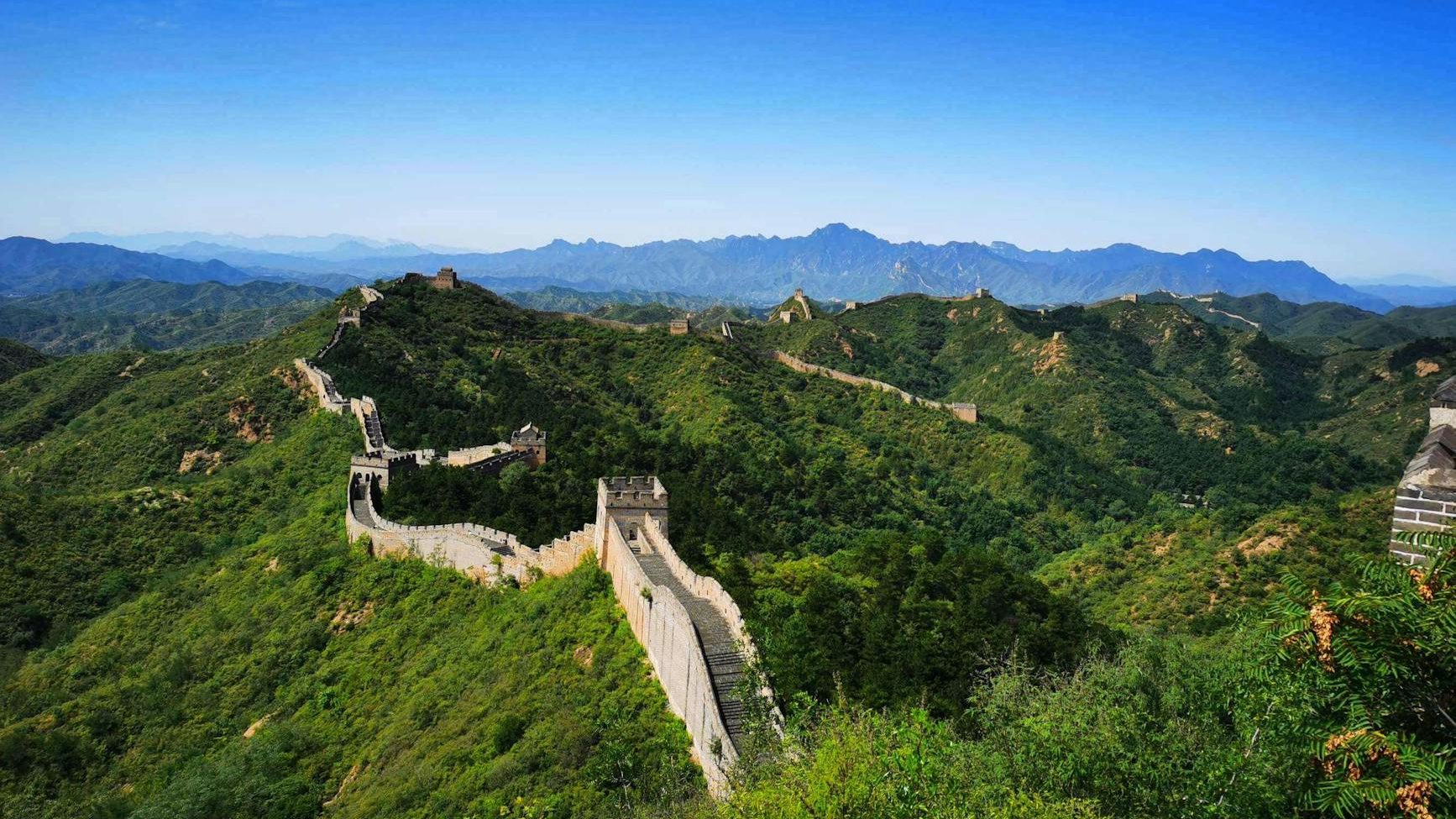 A panoramic view of the Great Wall of China winding through lush green hills under a bright blue sky.