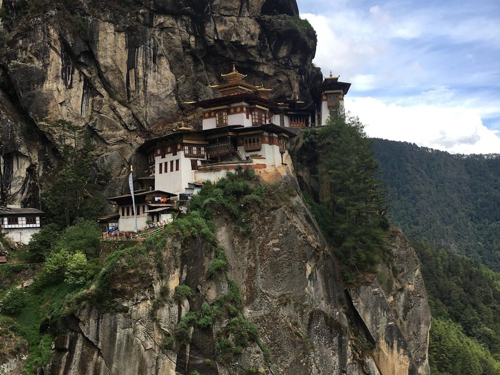 A monastery perched on a cliffside in Bhutan, surrounded by mountains and trees under a cloudy sky.