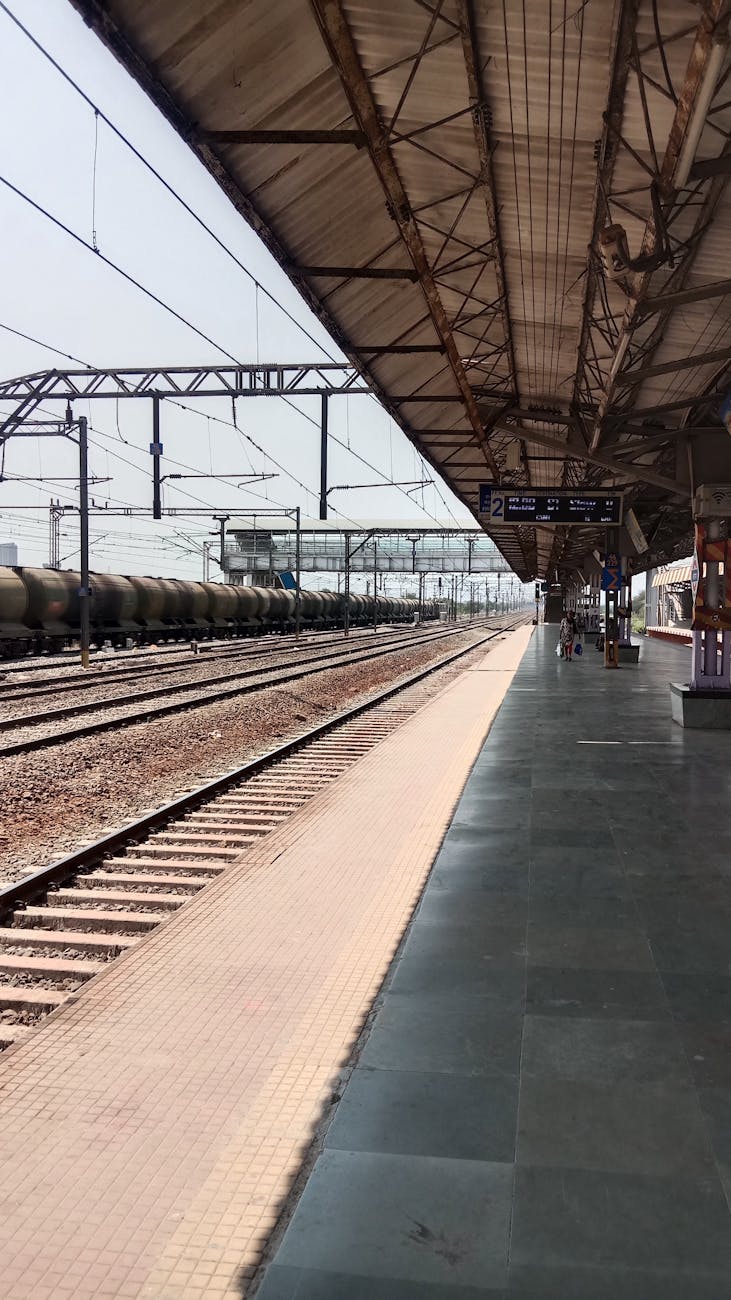 View of a train station platform with tracks, overhead structures, and a distant train in motion.