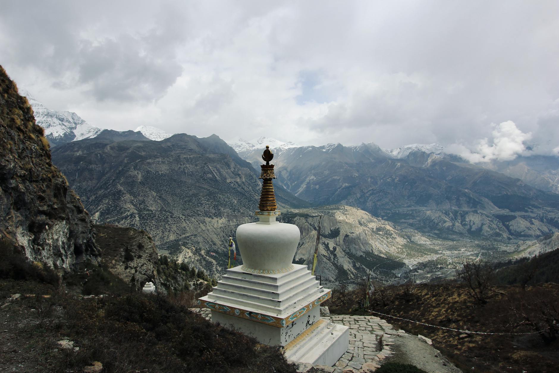 A white stupa sits on a mountainside, overlooking a vast landscape of rugged mountains and valleys under a cloudy sky.