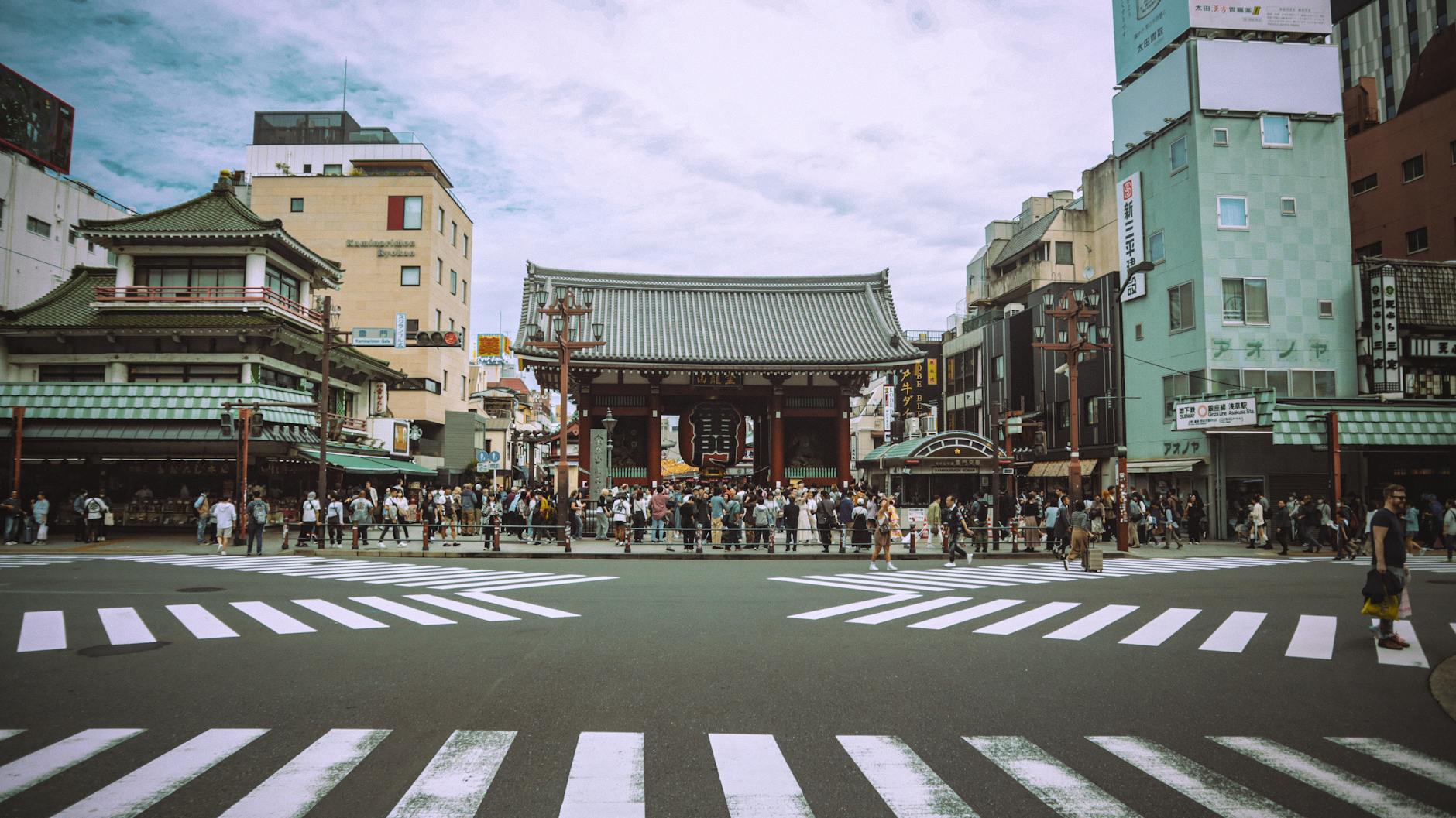 A busy street scene with a large traditional Japanese gate in the background, bustling with pedestrians, and modern buildings lining the street.