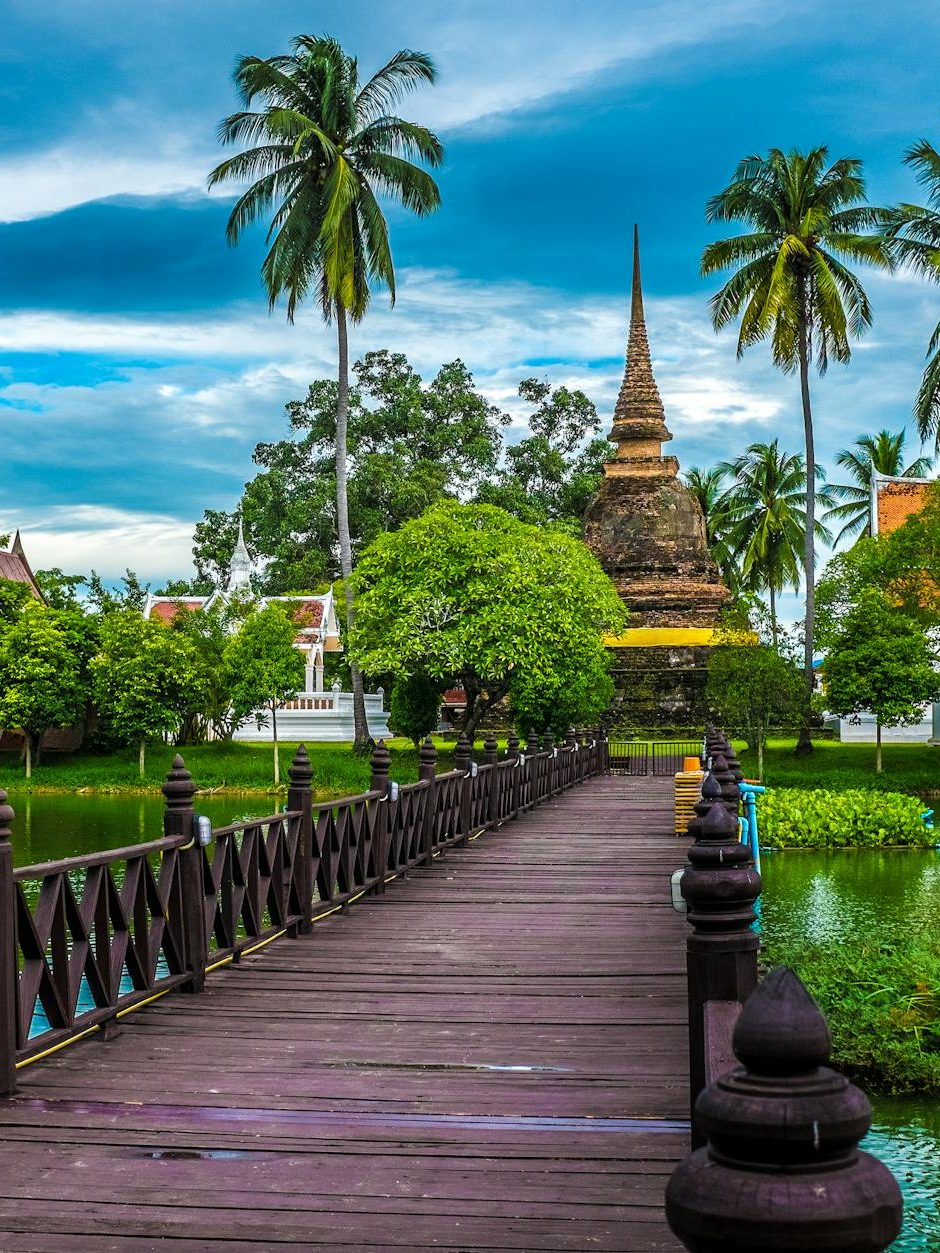 A wooden bridge leading to a historical temple surrounded by lush greenery and palm trees, with a serene water body in the foreground and dramatic clouds in the sky.