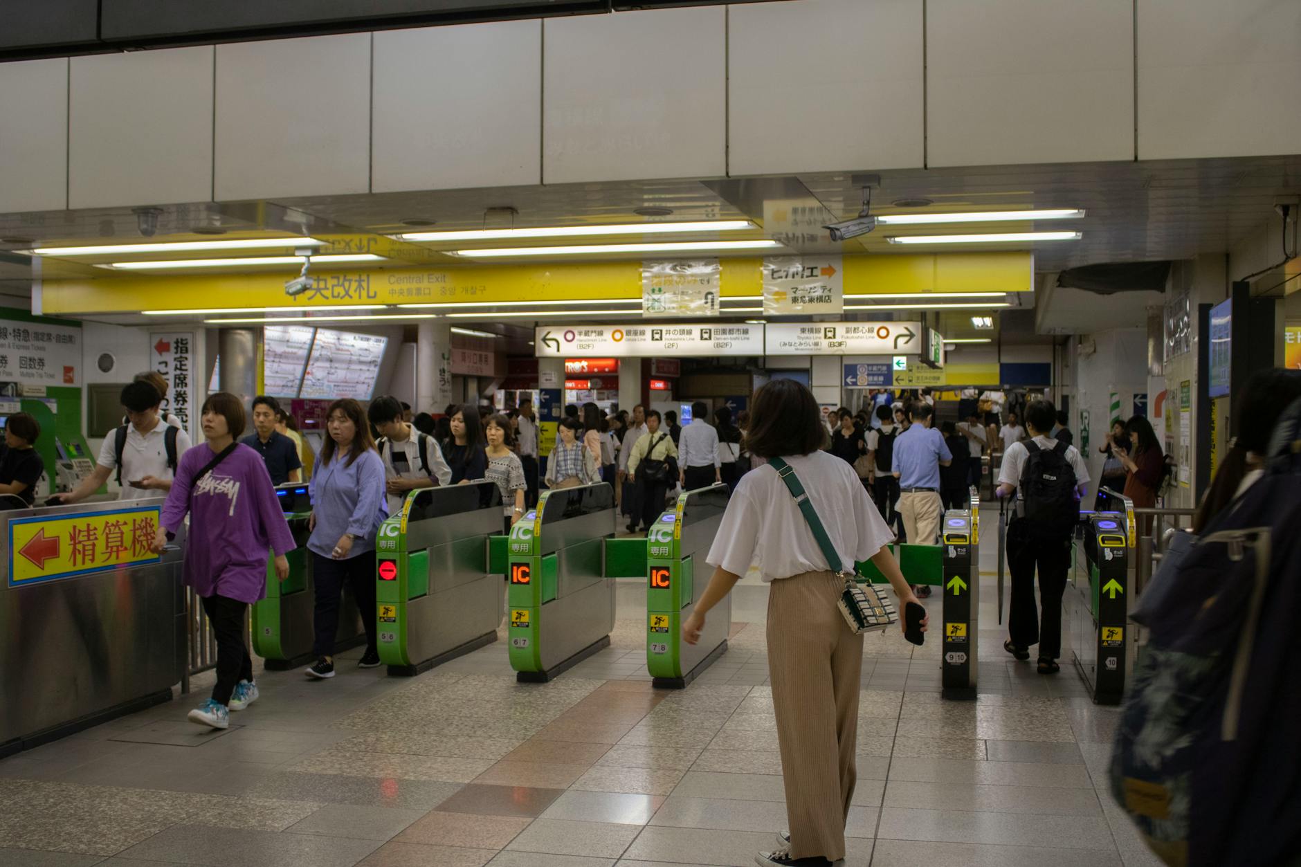 A busy subway station with numerous commuters exiting and entering through automated ticket gates. The environment is brightly lit, with signs in multiple languages and a large crowd of people moving in various directions.