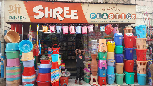 View of a plastic goods store named 'Shree Plastics', featuring an assortment of colorful buckets, crates, and bins stacked outside the shop. A person stands in front, appearing surprised or overwhelmed.